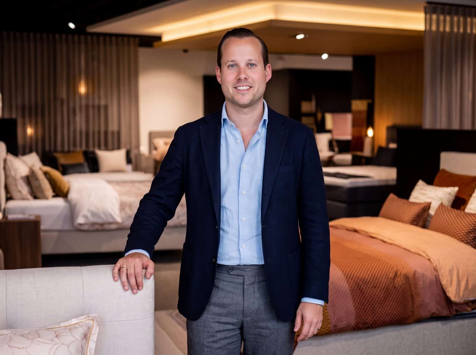 Adviseur van Grisar Slaapcomfort poseert in blazer in de toonzaal met opgemaakte bedden