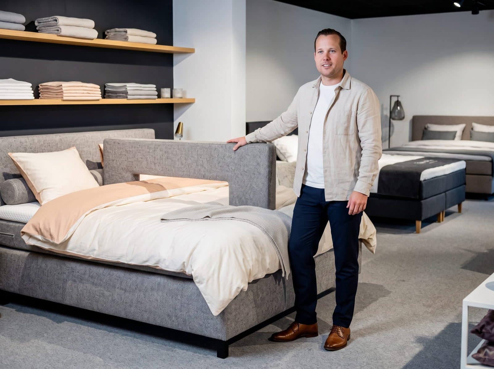 Adviseur van Grisar Slaapcomfort staat in de toonzaal naast een grijs boxspringbed met beddengoed