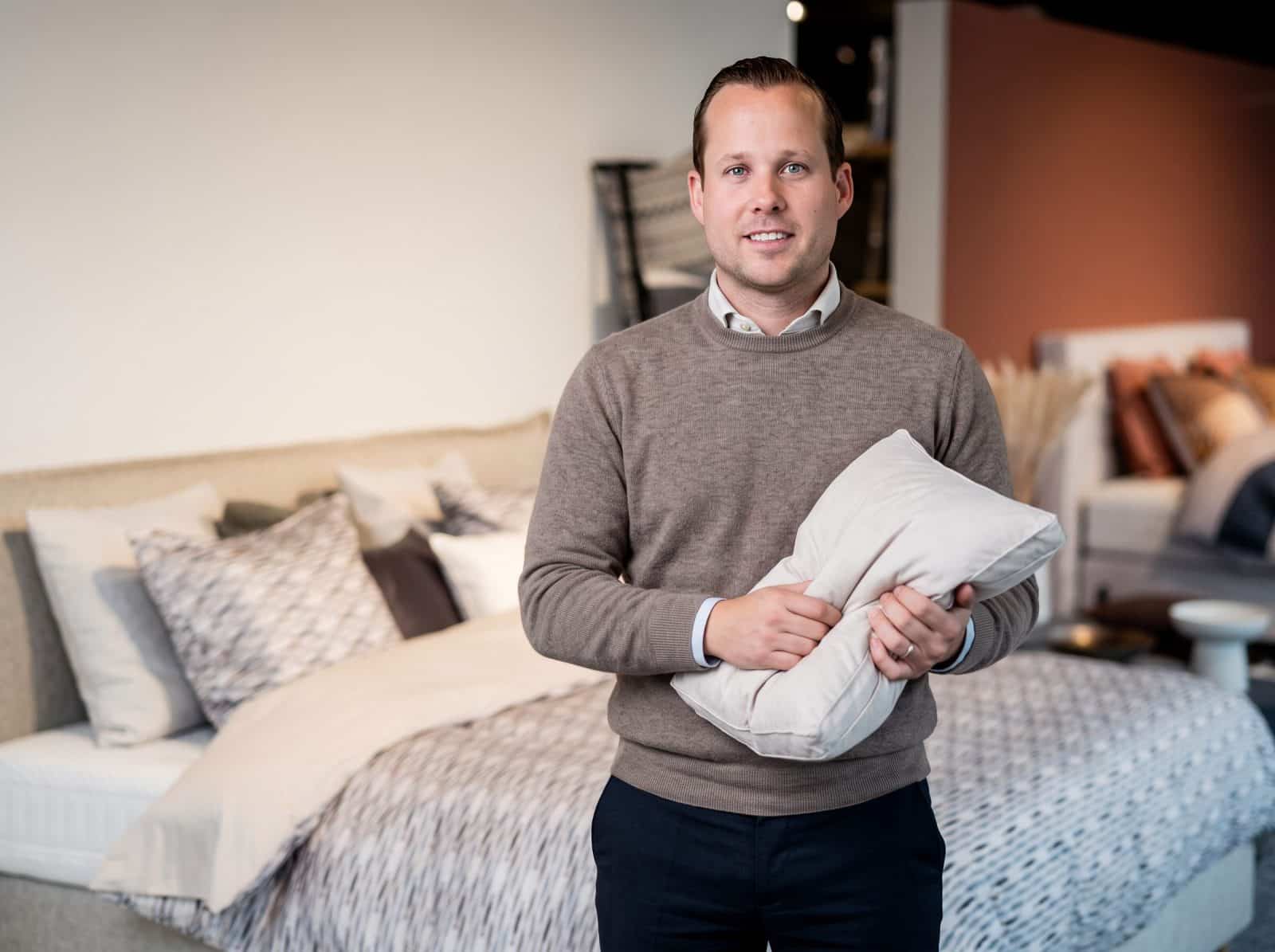 Adviseur van Grisar Slaapcomfort houdt een hoofdkussen vast voor een opgemaakt bed in de toonzaal