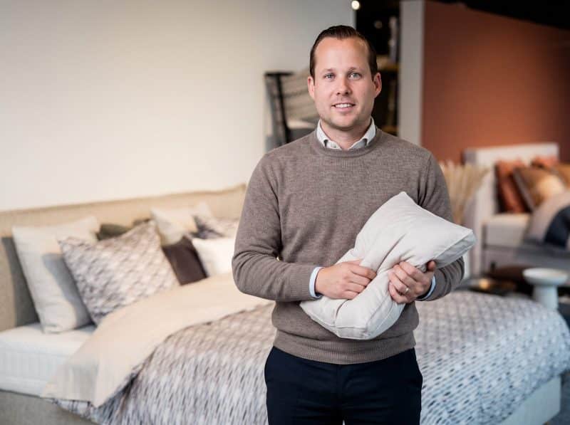 Adviseur van Grisar Slaapcomfort houdt een hoofdkussen vast voor een opgemaakt bed in de toonzaal
