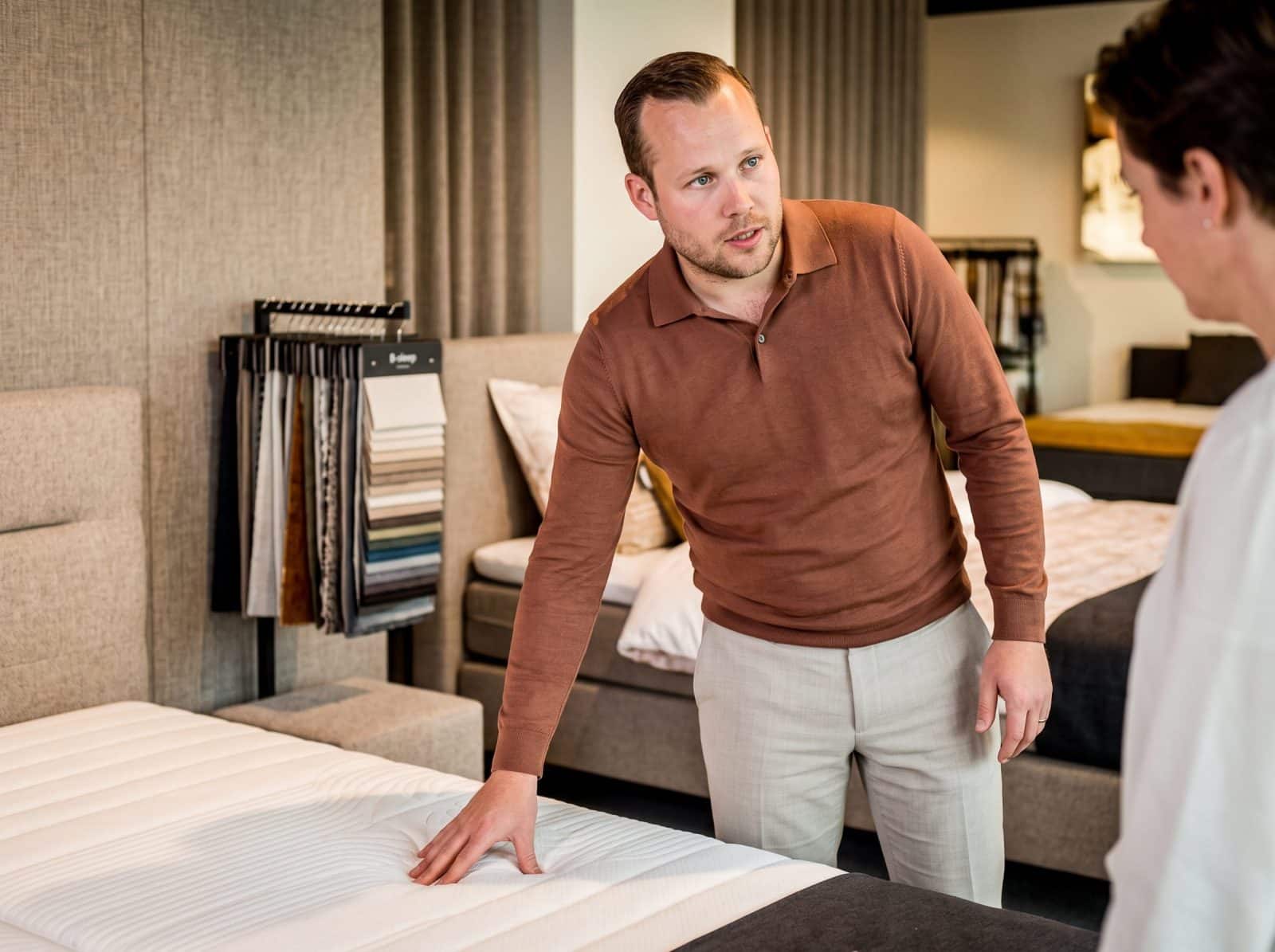 Adviseur in bruine trui voelt aan een matras bij een stalenrek in de toonzaal