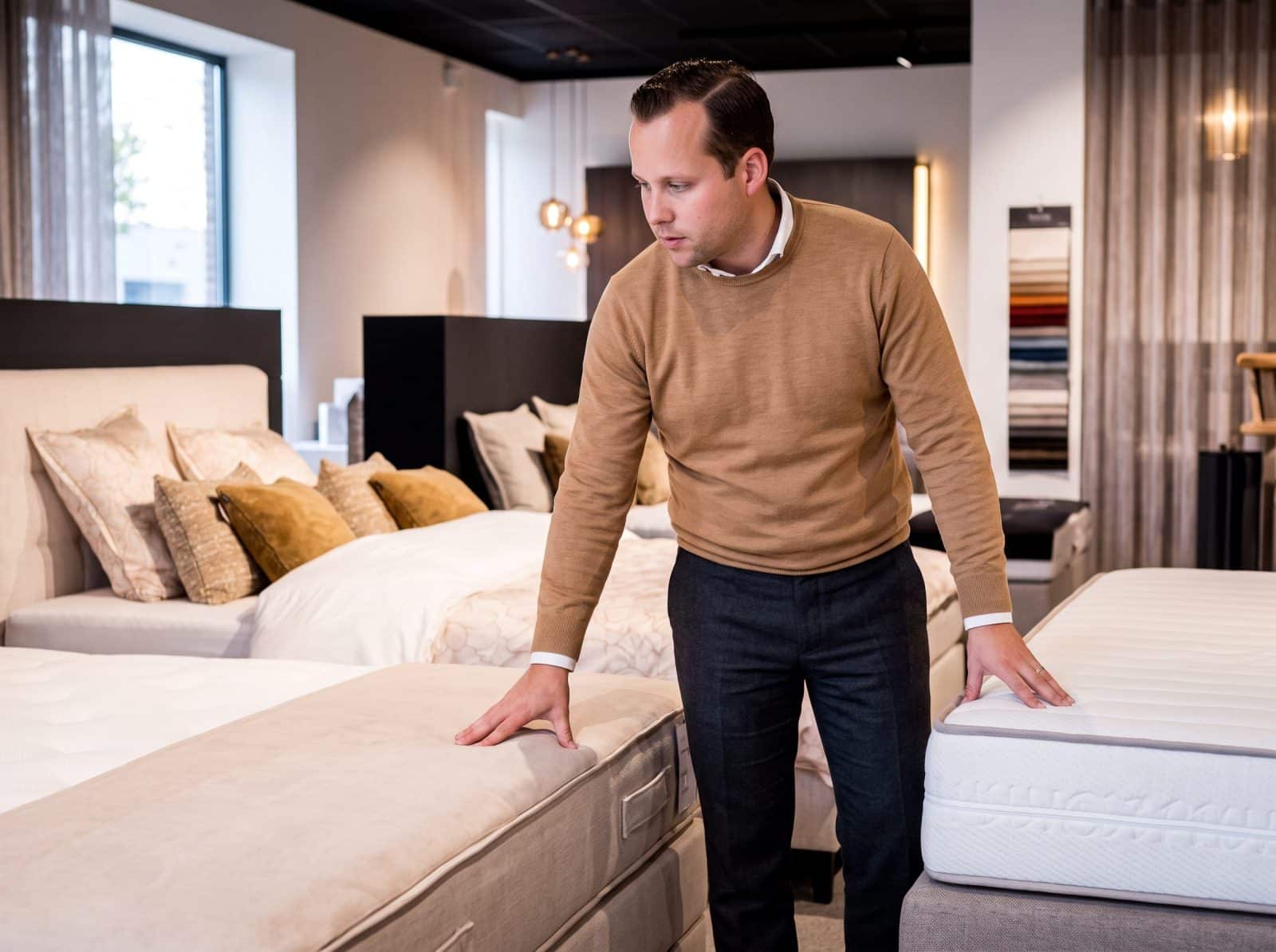 Adviseur in een bruine trui leunt over een matras en een bed in de toonzaal