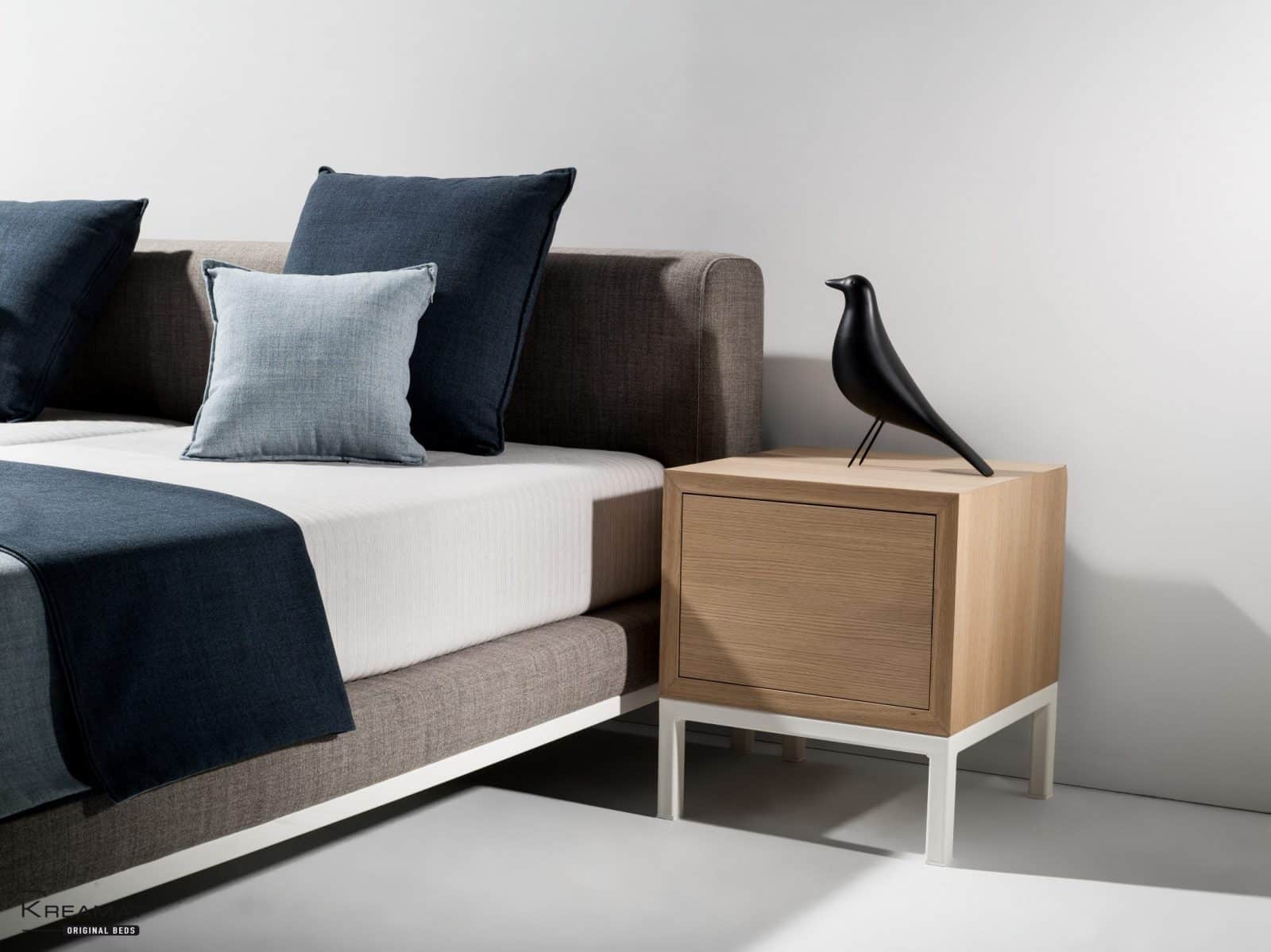 Bed met grijze bedbodem, witte matras en donkerblauwe kussens naast een houten nachtkastje
