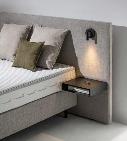 Grijs gestoffeerd hoofdbord met leeslamp en kussens, naast een matras