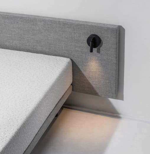 Close-up van een matras tegen een grijs gestoffeerd hoofdbord met leeslamp