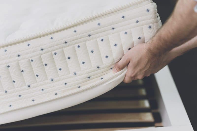 Hand tilt de rand van een witte matras op, waardoor het pocketveringsysteem en de lattenbodem zichtbaar zijn