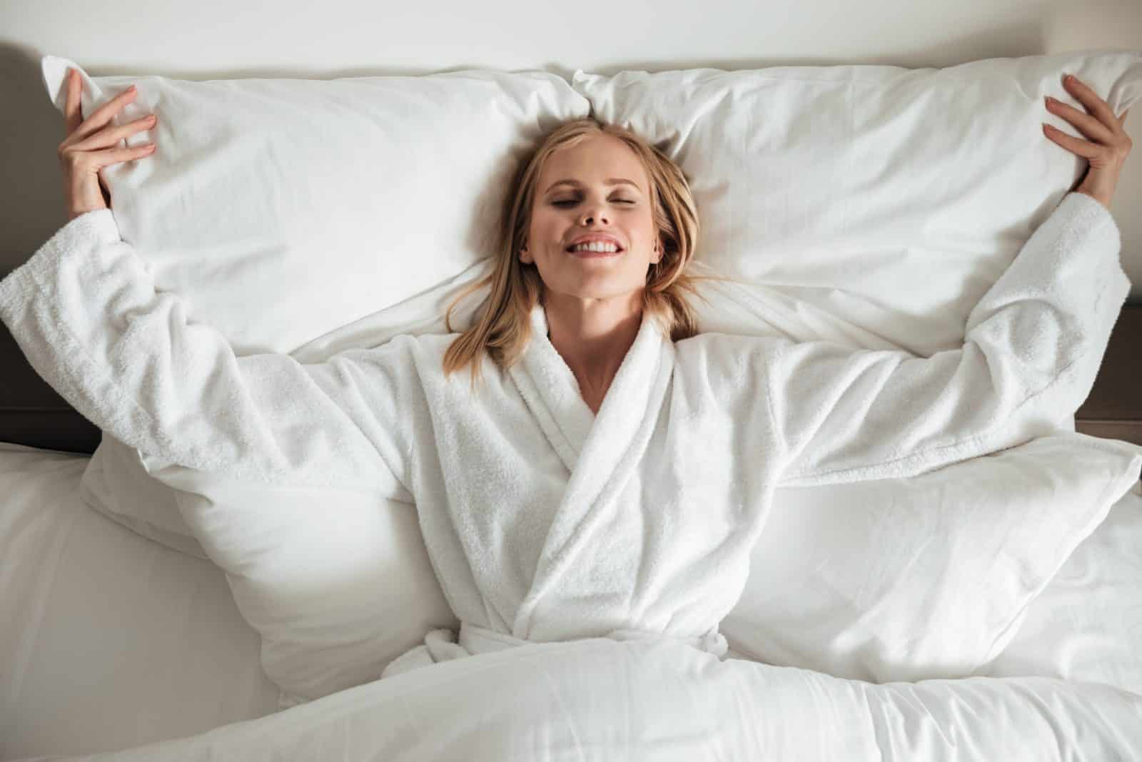 Lachende vrouw in een witte badjas die op een wit hoofdkussen en dekbed ligt
